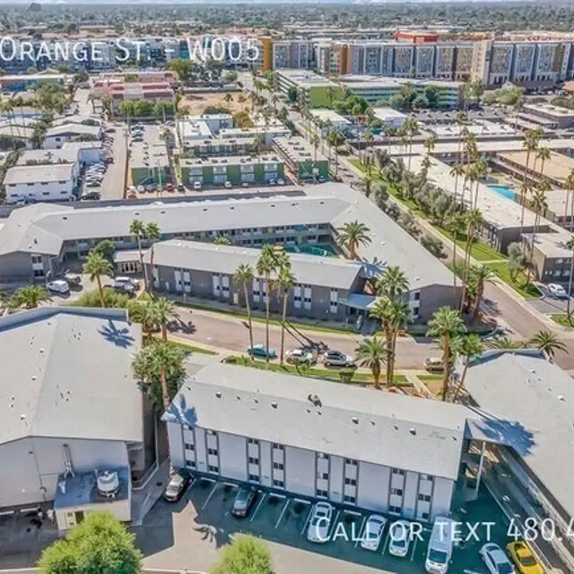 Aerial view of an apartment complex in a suburban area, showing multiple buildings, palm trees, and parked cars, with a cityscape in the background.