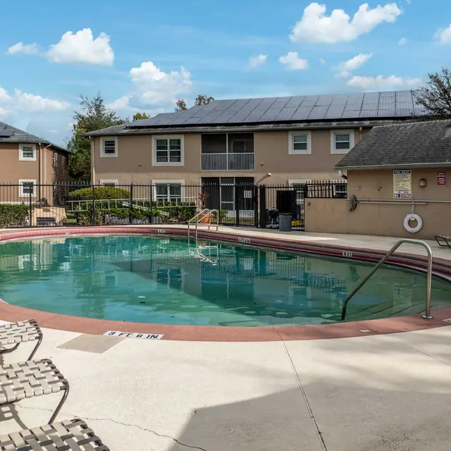 A swimming pool surrounded by lounge chairs, with a fence in the background and apartments overlooking the area.