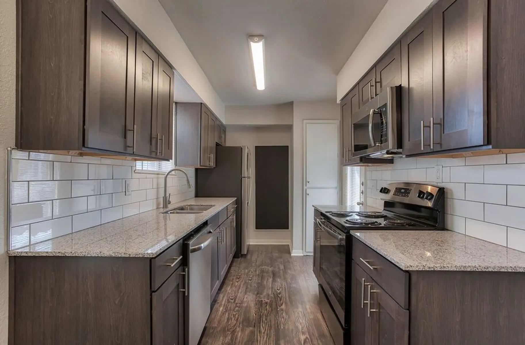 A modern kitchen featuring dark wooden cabinets, granite countertops, and stainless steel appliances. The kitchen has a spacious layout and includes a sink, oven, and refrigerator, with white subway tile backsplash.