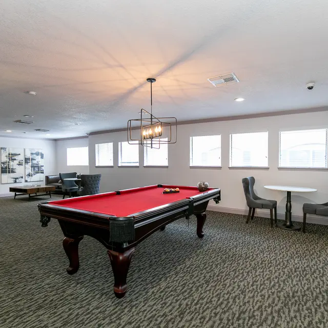 A spacious game room featuring a red felt pool table in the center, surrounded by plush seating and a dining table. Large windows allow natural light to fill the room, which has a modern decor with carpeted flooring.