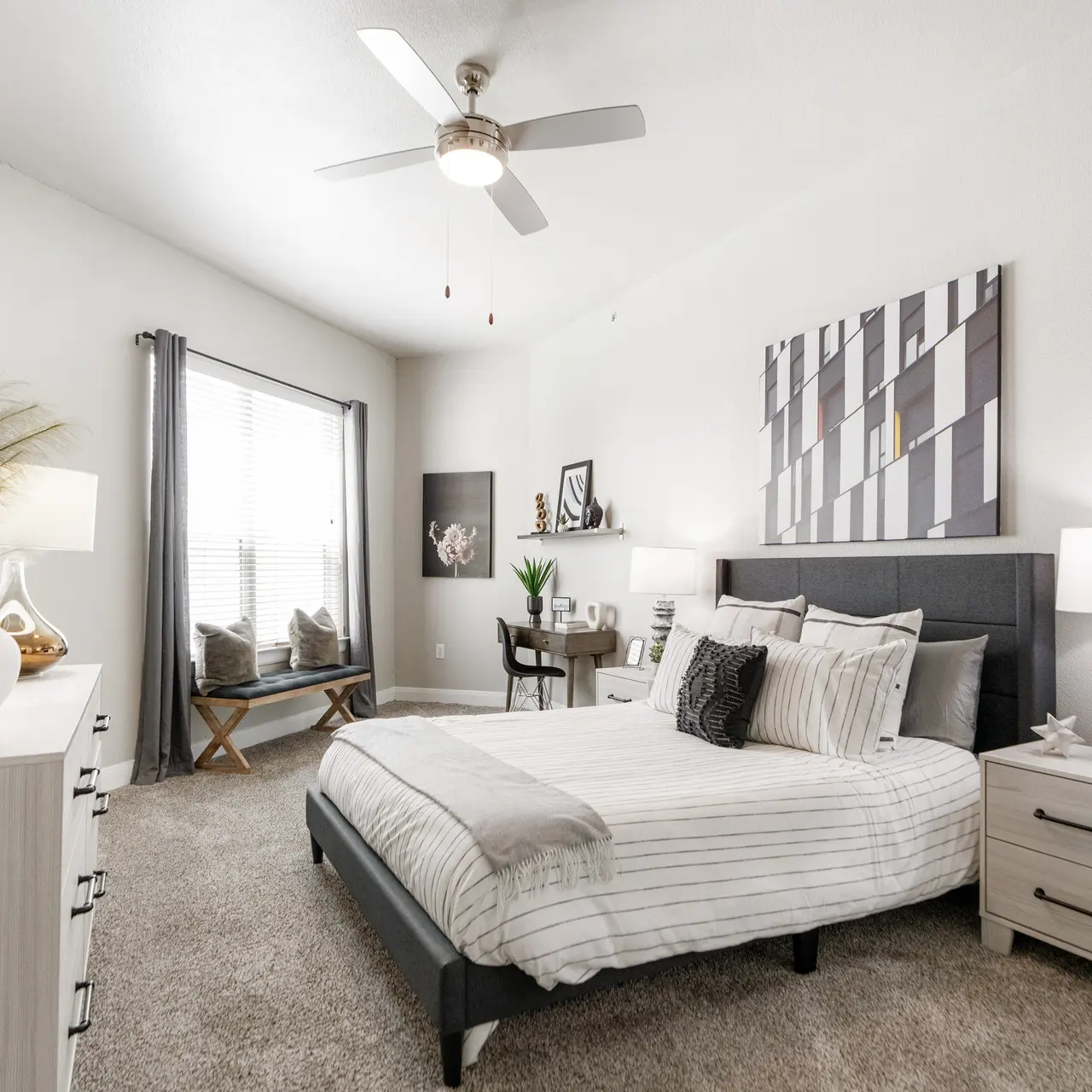 A modern bedroom featuring a large bed with striped bedding, two bedside tables with lamps, and a window with gray curtains. A dresser and a small desk with chairs are also visible, along with decorative elements including a plant and artwork.