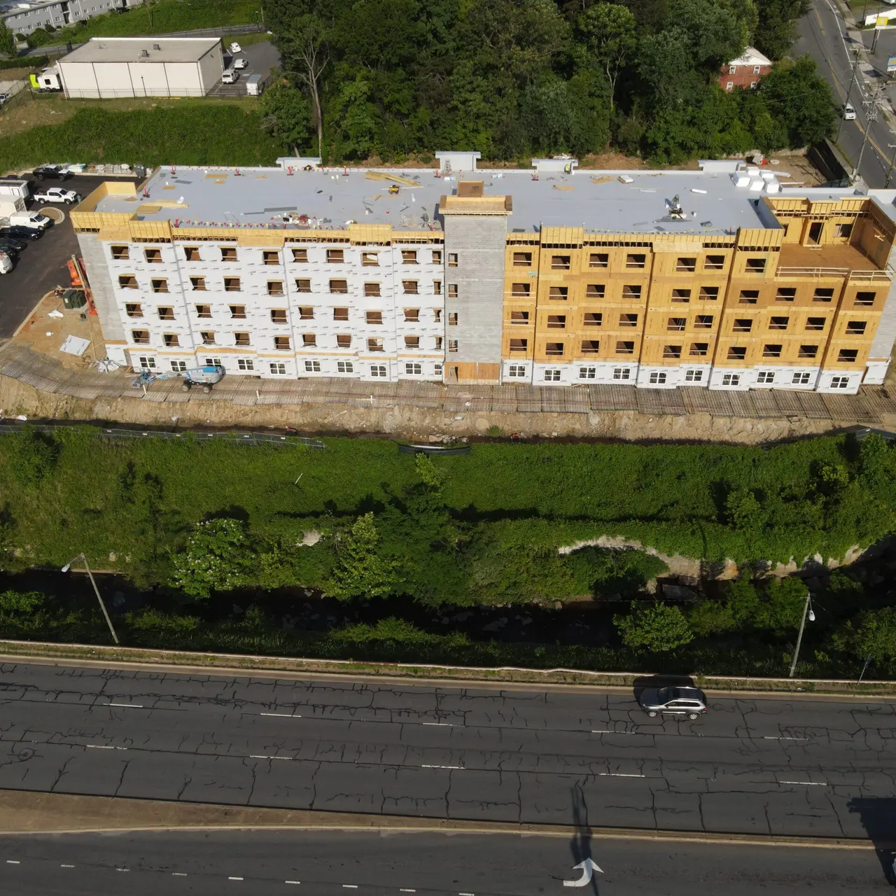 Aerial view of a new multi-story building under construction, featuring a mix of wood and white siding, set above a green area adjacent to a road.