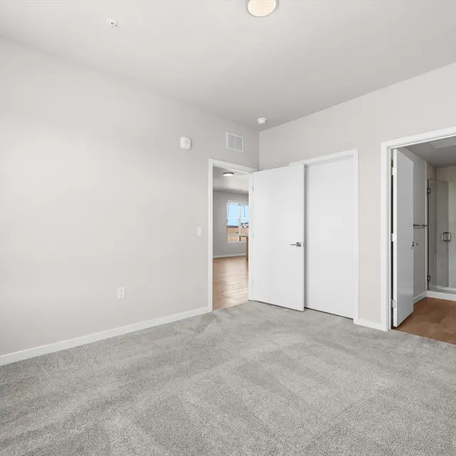 A spacious empty room with light gray carpet and neutral-colored walls. The room features two open white doors leading to other areas, and a window can be seen in the background providing natural light.