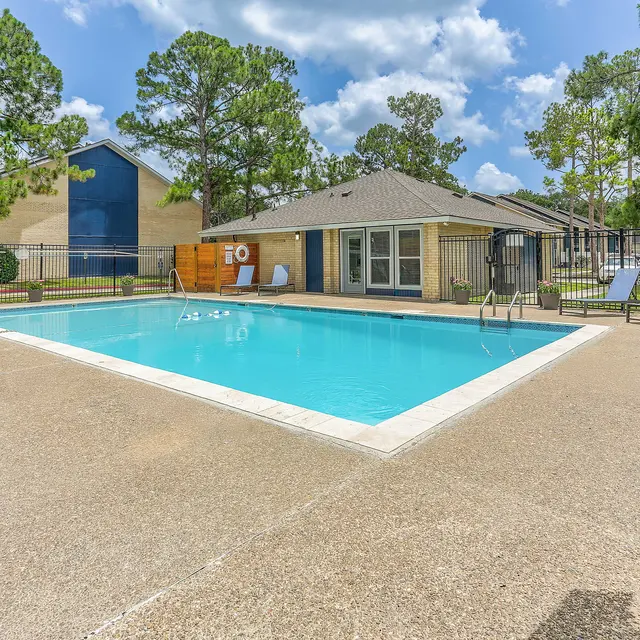 A clear swimming pool surrounded by lounge chairs and greenery, with a building in the background under a blue sky.