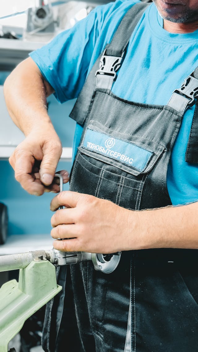 A mechanic in a blue shirt and overalls is working with a tool on a machine. The mechanic is focused on his task, with a workspace in the background that includes various tools and equipment.