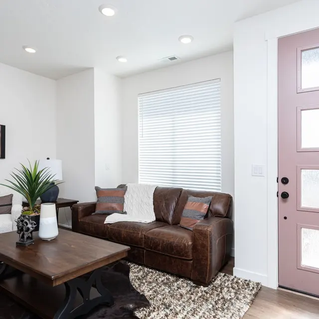 A modern living room featuring a brown leather sofa, a white armchair, and a wooden coffee table with decorative items. A pink front door is visible along with a large window with white blinds.