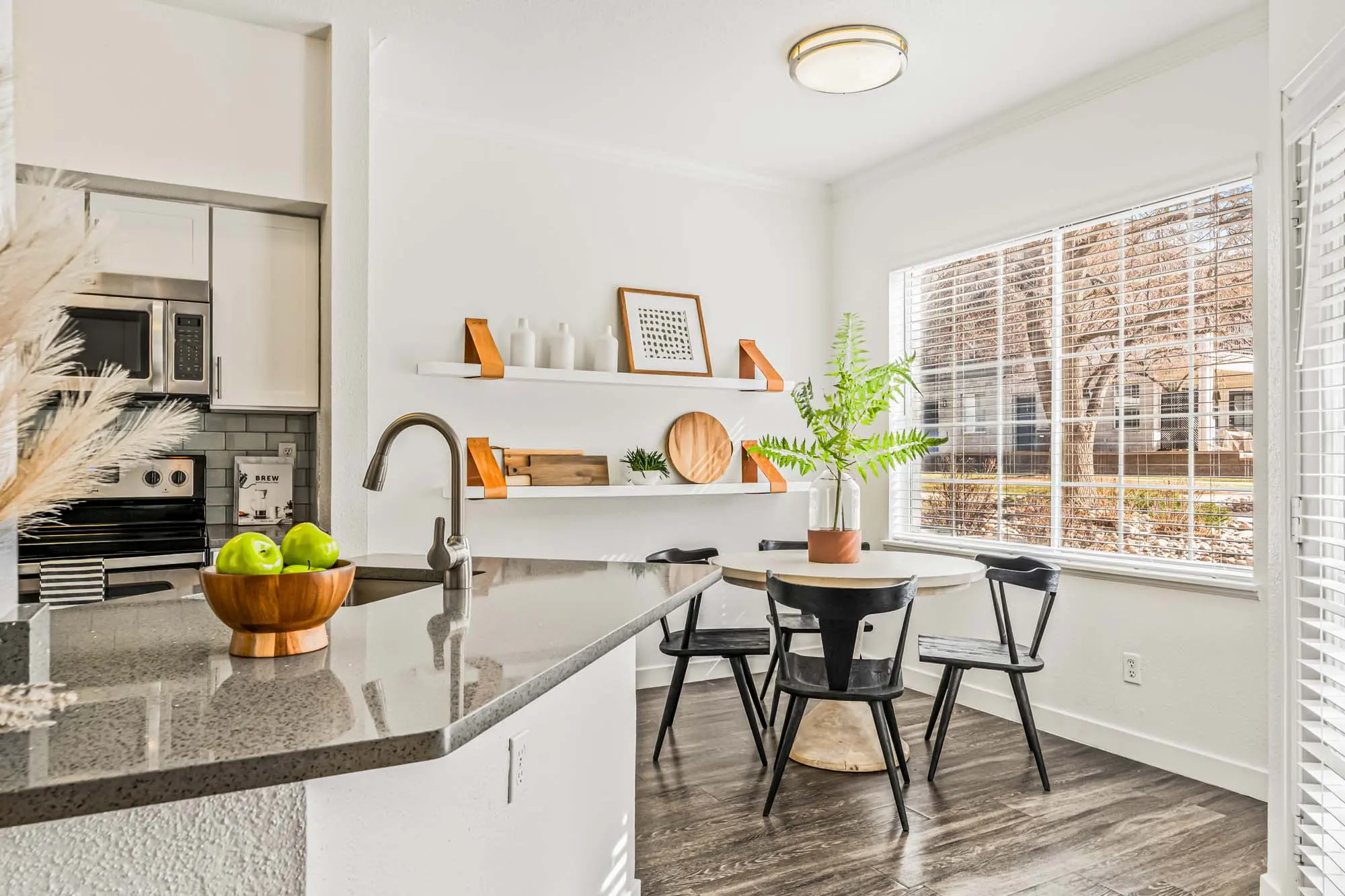 A bright kitchen and dining area featuring a round table with black chairs, a potted plant, and open shelving with decorative items.