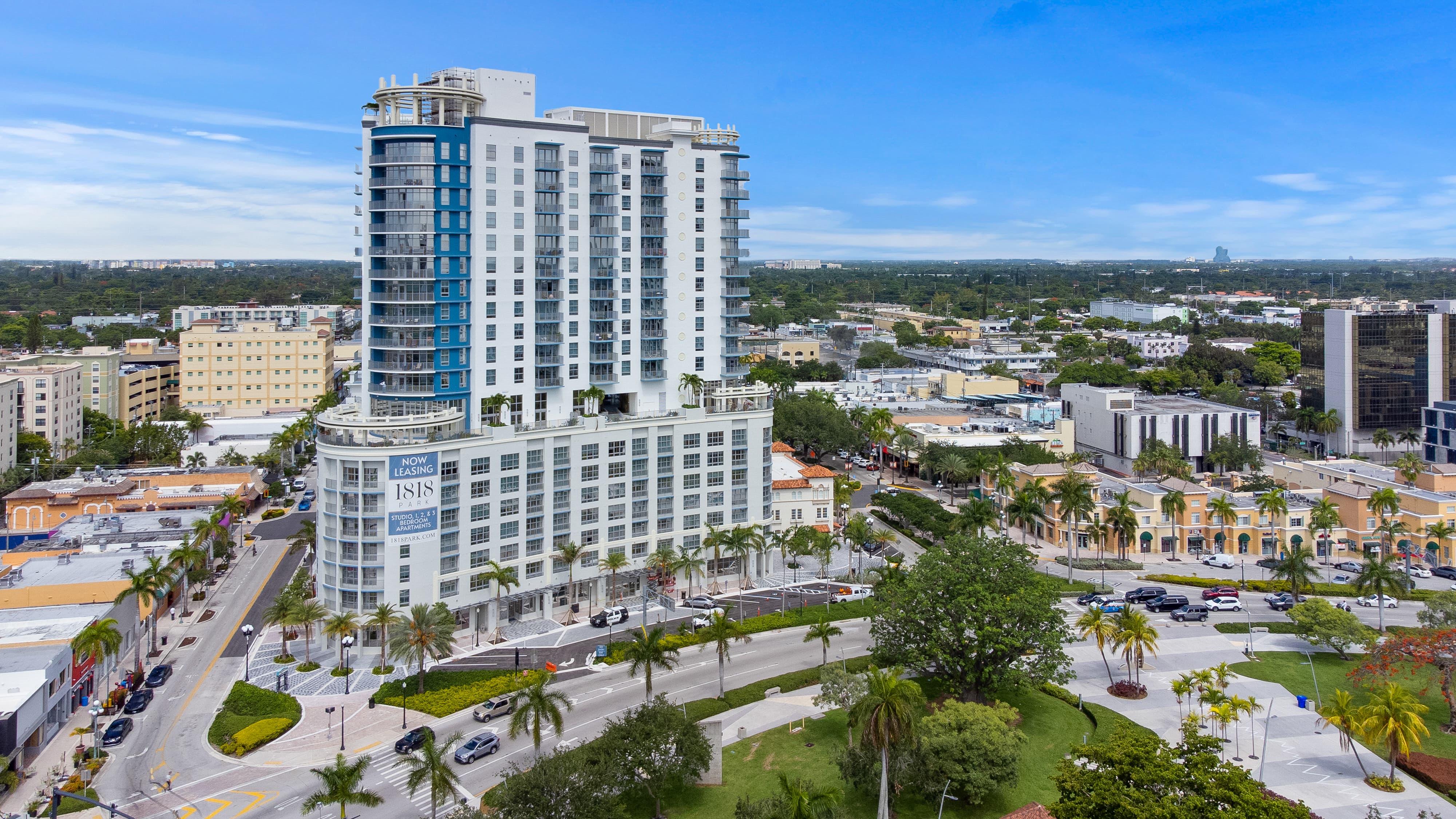 A modern high-rise building with a glass and concrete facade in an urban area, surrounded by palm trees and other buildings.