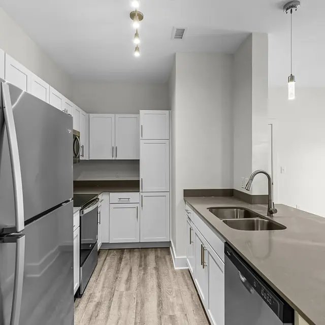 A modern kitchen featuring stainless steel appliances, including a fridge, microwave, and dishwasher, with white cabinetry and a dark countertop. The kitchen is illuminated by pendant lights.