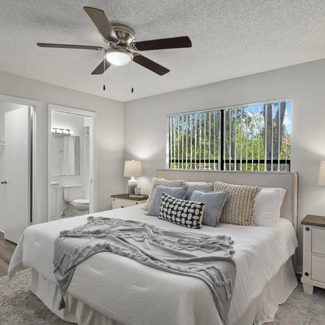 A cozy bedroom featuring a queen-sized bed with decorative pillows, a nightstand with a lamp, and a ceiling fan. A window allows natural light, and a bathroom is visible through a door on the left.