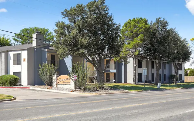 Exterior view of Sunset Apartments featuring three floors, landscaped greenery, and trees, along a street on a sunny day.