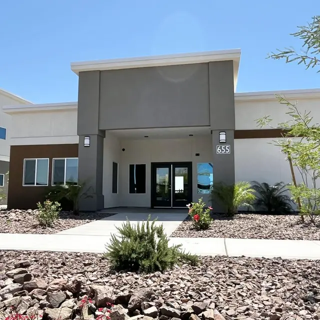 Modern Building Entrance A modern building with a prominent entrance, featuring a combination of grey and brown exterior colors. The entrance is framed with large glass doors, and the surrounding landscape includes small bushes and rocks under a clear blue sky.