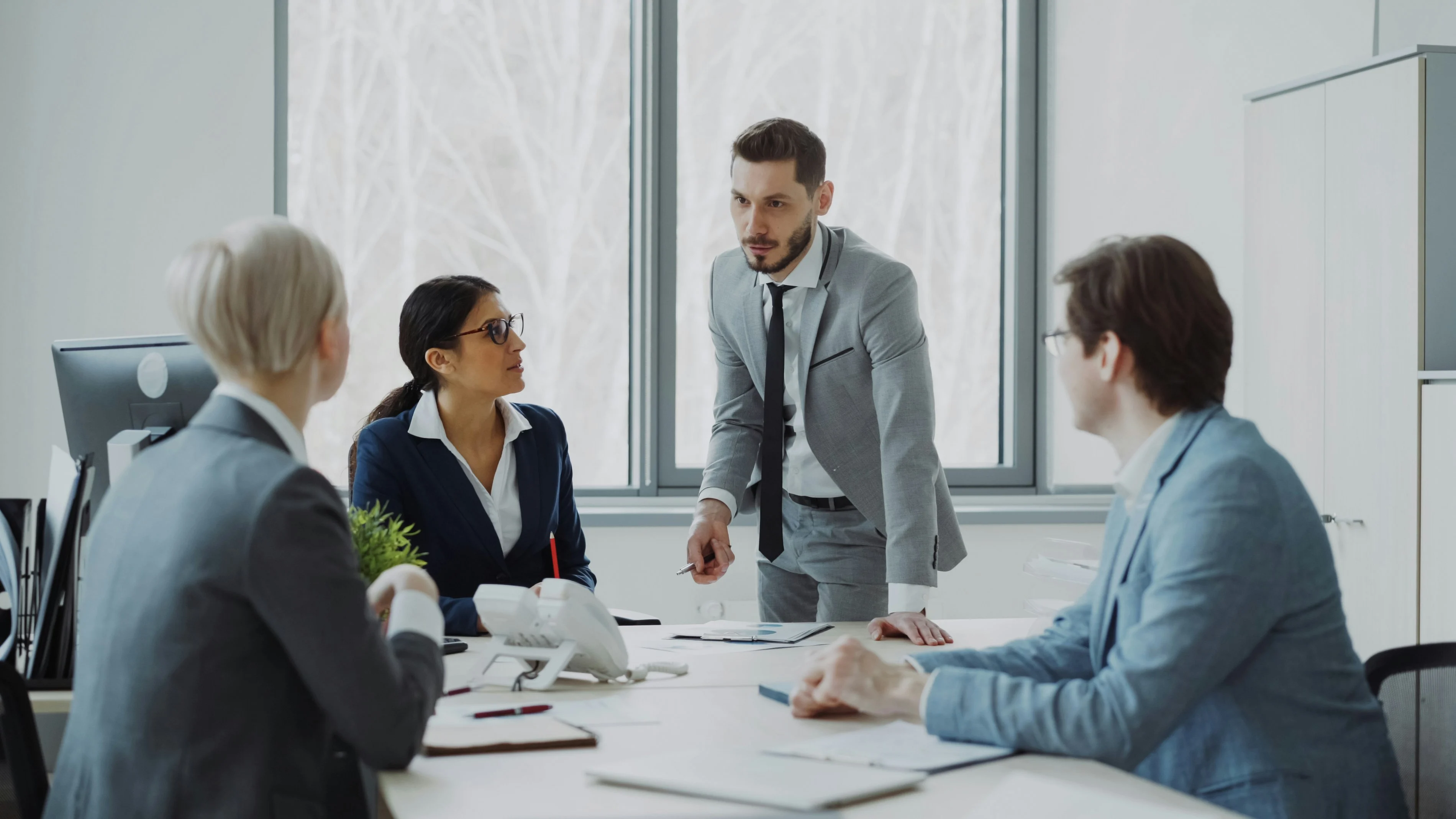 Professional Business Meeting A business meeting in a modern office setting. Four professionals are seated around a table, engaging in discussion. One man in a suit stands while addressing the seated team, which includes a woman in glasses and a woman with short white hair. Large windows provide a view of trees outside.