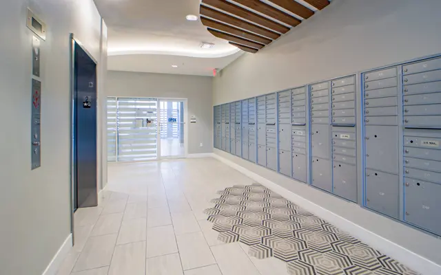 A modern, well-lit mailroom featuring multiple mailboxes along one wall, adjacent to an elevator and a glass door leading outside. The tile floor has a geometric pattern, and the ceiling is designed with wooden slats.