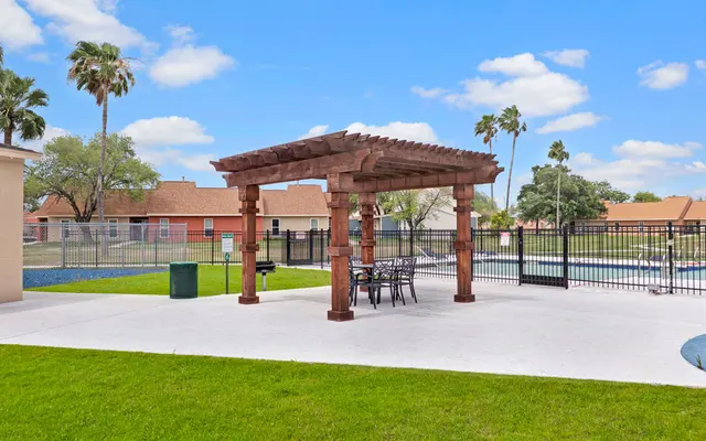 Outdoor Patio with Pergola A pergola with dining furniture in an outdoor area surrounded by greenery and a pool.