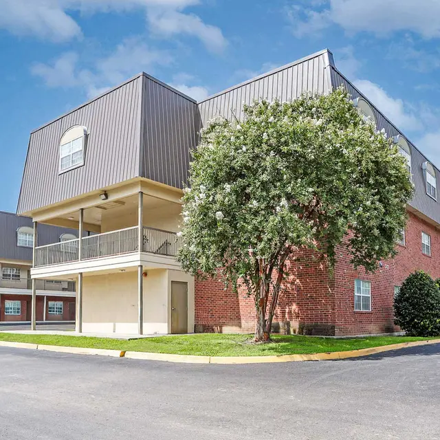 A modern multi-story building with a combination of brick and metal exterior, surrounded by trees and landscaped grass areas, under a blue sky with clouds.