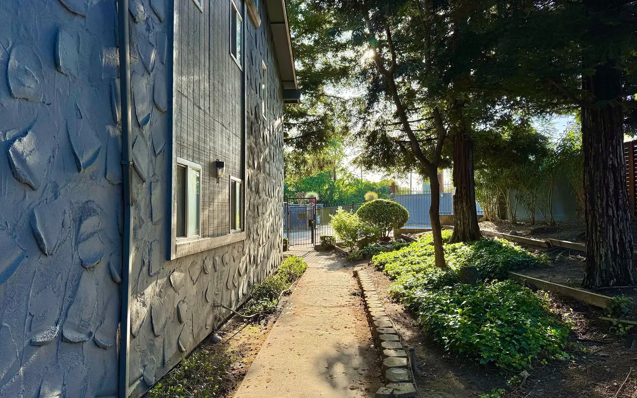 A walkway leading beside a building, bordered by lush greenery and trees, with a clear blue sky in the background.