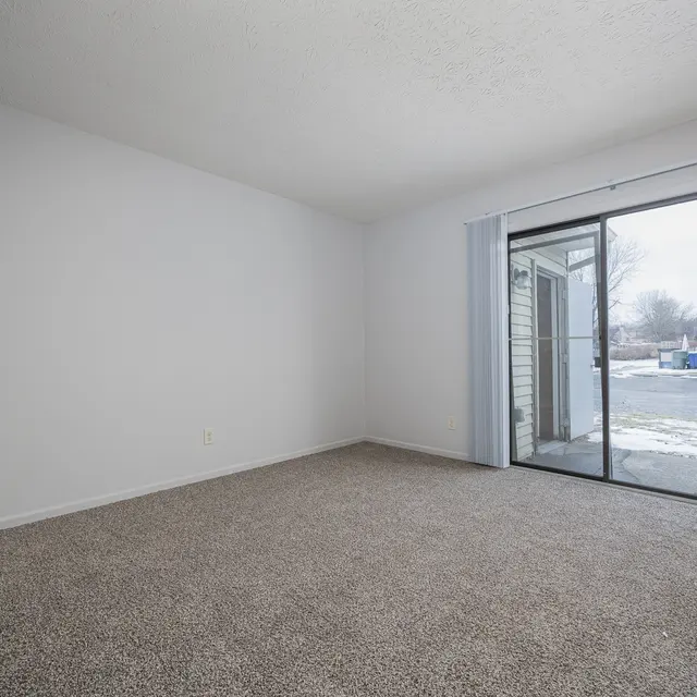 An empty room with light gray walls and a beige carpet. There is a sliding glass door leading to an outdoor area, allowing natural light to enter the room.