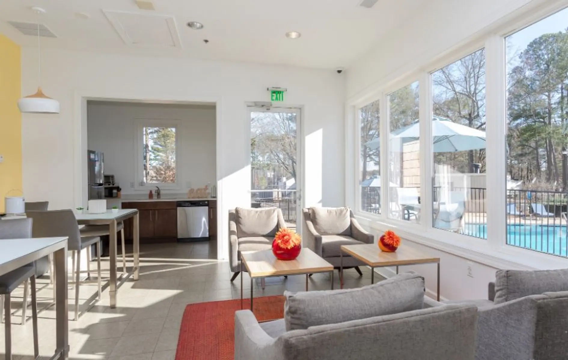 A bright and modern living area featuring comfortable seating, a dining table, and large windows overlooking a pool area. Decorated with orange flowers on the tables, the space is filled with natural light and has a warm atmosphere.