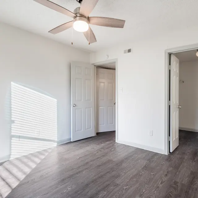 An empty room featuring light-colored walls and a ceiling fan, with two closed doors on opposite walls. Sunlight casts shadows on the floor from the window.
