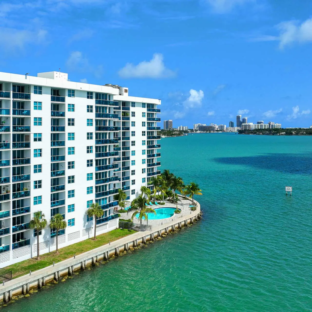 View of a waterfront apartment building next to a turquoise body of water with a small swimming pool and city skyline in the distance