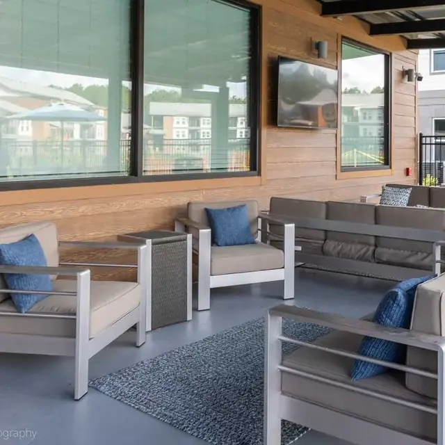 An outdoor lounge area featuring modern seating with light-colored cushions and blue decorative pillows. The background has large windows overlooking a fenced yard. A flat-screen TV is mounted on the wall.