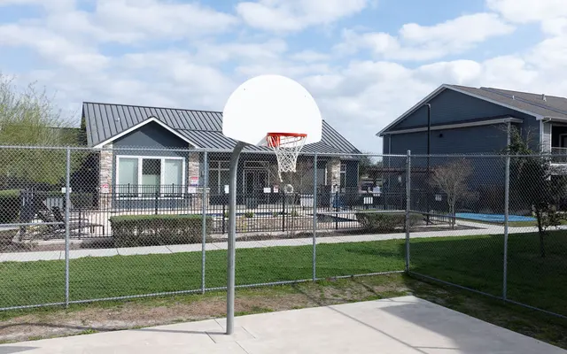 A fenced basketball court with a hoop in front of residential buildings and a swimming pool area.