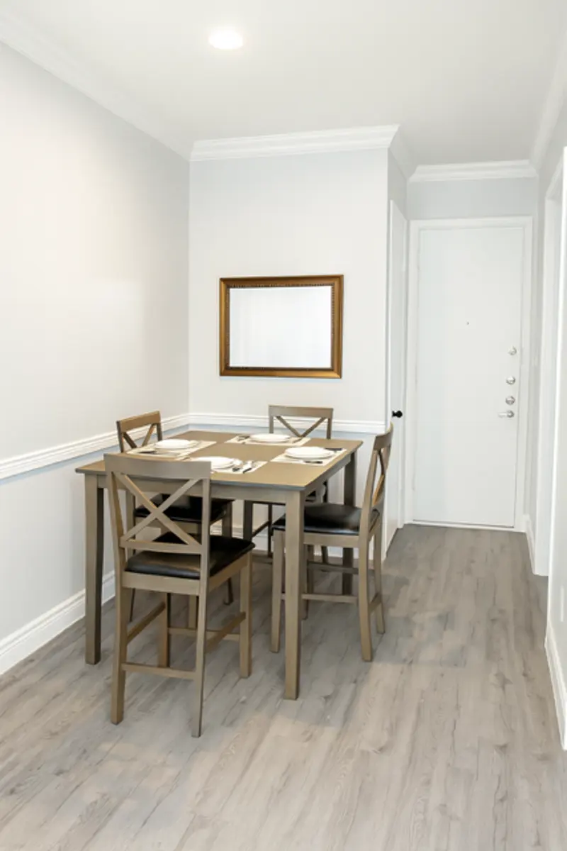 A small dining area with a wooden table and four chairs, light wooden flooring, and a wall-mounted mirror.