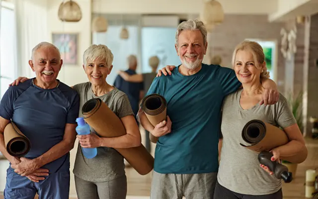 Seniors Embracing Fitness Together A group of four seniors standing together in a yoga studio, smiling happily while holding yoga mats and a water bottle.