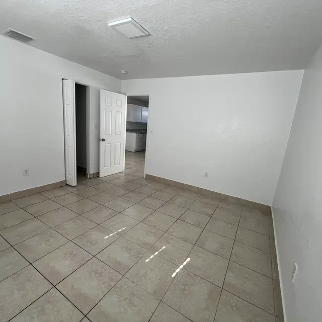 Empty room with tiled floor, white walls, and a doorway leading to another room. Natural light enters through a window.