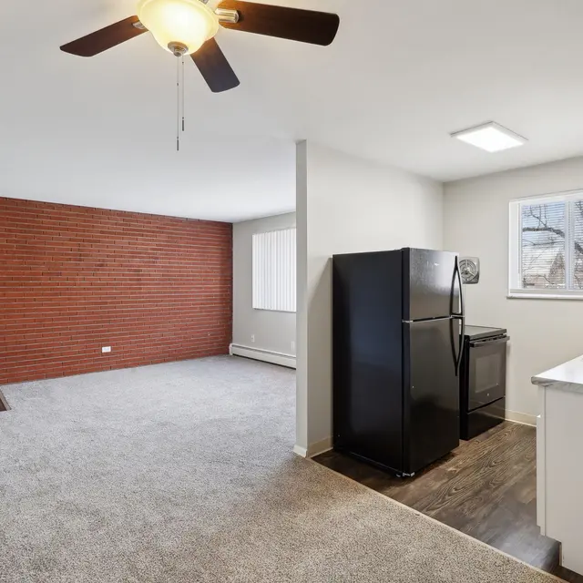 Interior view of a modern apartment featuring a brick accent wall, carpeted living area, and a kitchen area with a refrigerator and sink.