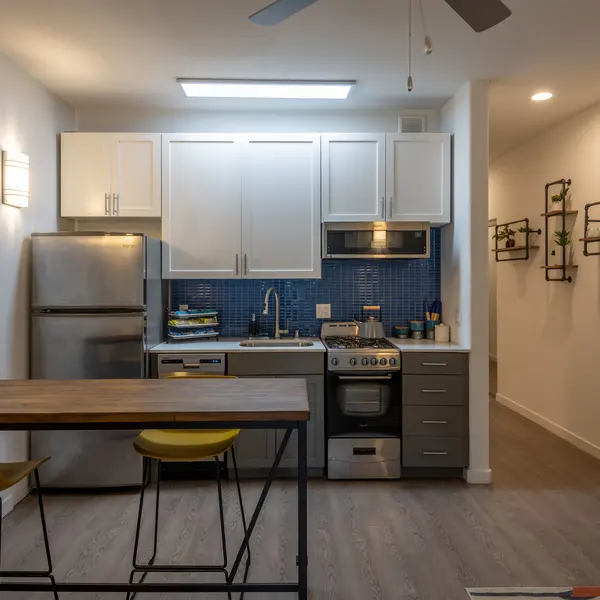 A modern kitchen featuring white cabinets, a stainless steel refrigerator, a gas stove, and blue tiled backsplash, with a dining area that includes a wooden table and yellow chairs.