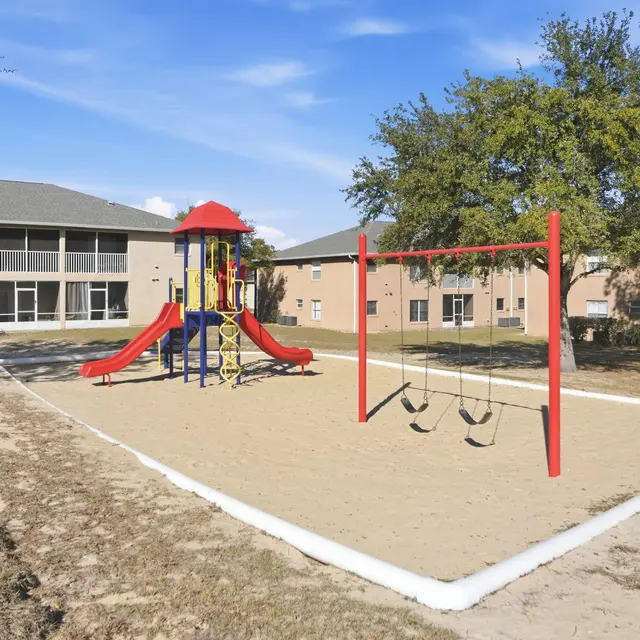 A playground featuring a colorful play structure with a slide, swings, and surrounding grassy space, set in front of residential buildings.