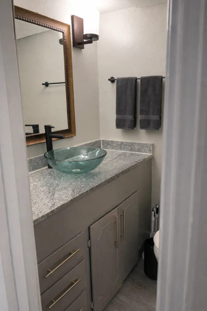 A modern bathroom featuring a gray countertop, a glass bowl sink, and a round mirror. The decor includes two gray towels hanging on a towel rack and a cabinet under the sink.