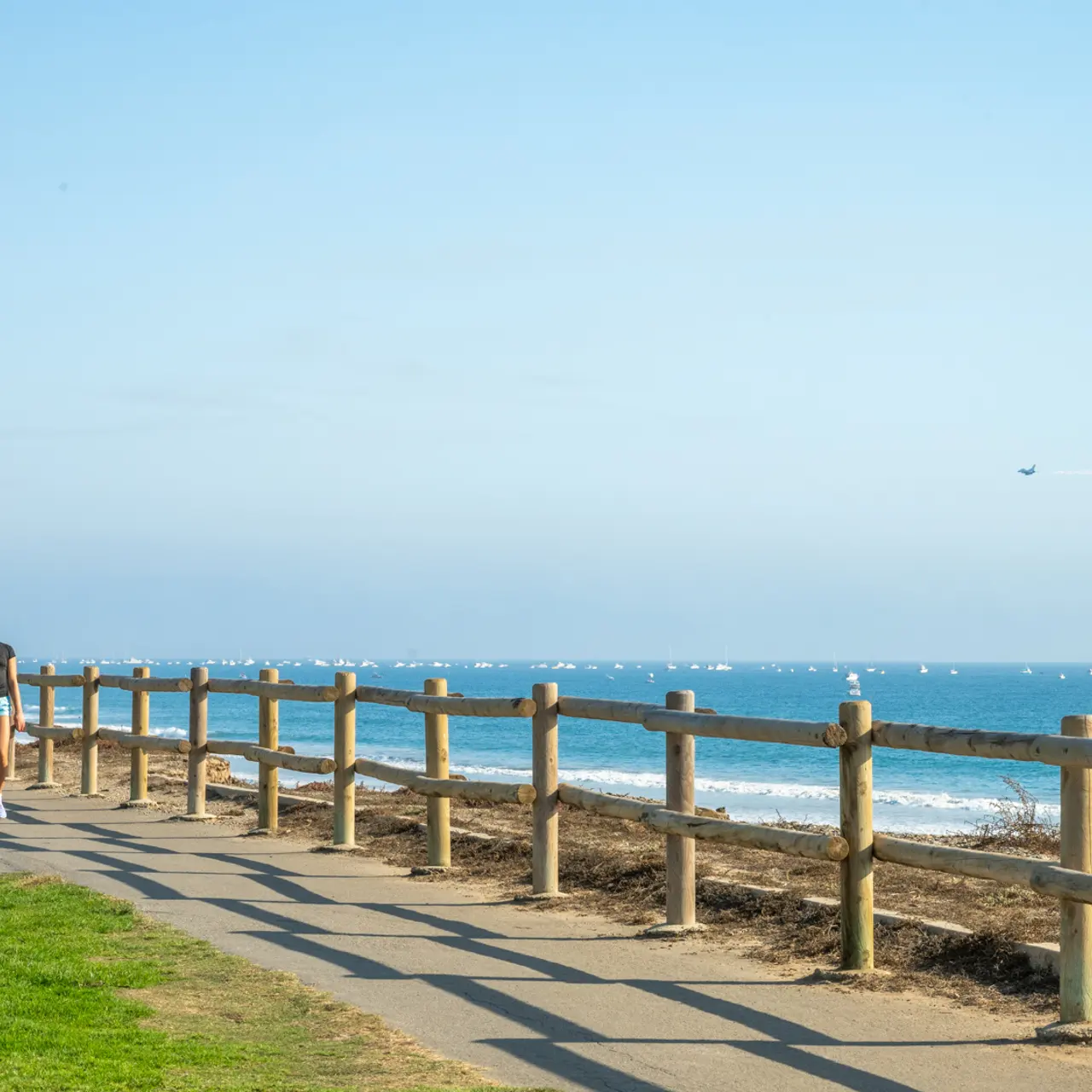 Two people walking along a coastal path near the ocean, with a wooden fence and green grass nearby.