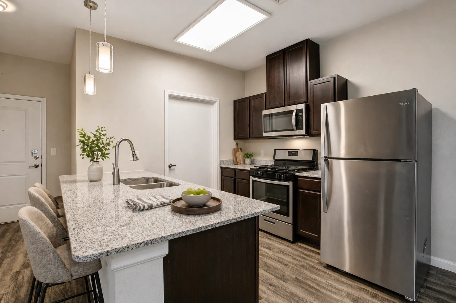 Modern Kitchen Interior A modern kitchen with dark wooden cabinets, stainless steel appliances, and granite countertops. The kitchen island has a small bowl of fruit and is accompanied by bar stools. A door leads to another room, and ceiling lights provide illumination.