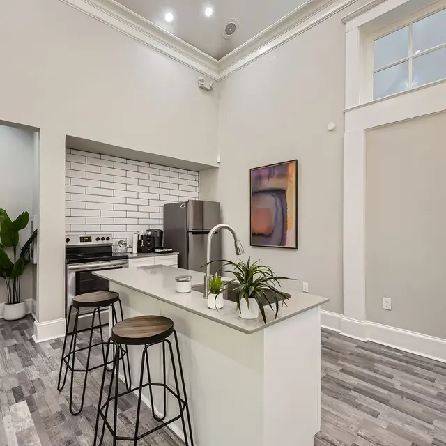 A modern kitchen with a minimalistic design featuring a kitchen island, bar stools, and stainless steel appliances. The walls are painted light gray, and there is a large window allowing natural light to enter.