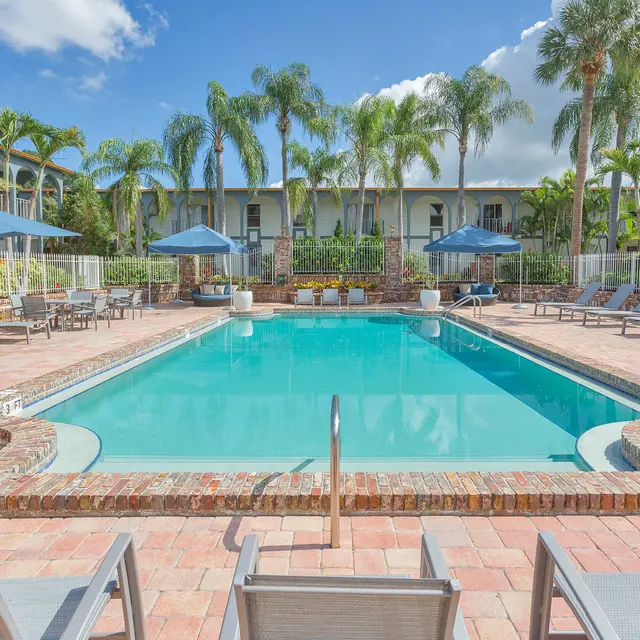 A bright outdoor swimming pool area surrounded by palm trees and sun loungers. The pool is clear with a brick border, and there are shaded seating areas beneath blue umbrellas. The backdrop features a multi-story building with green landscaping.