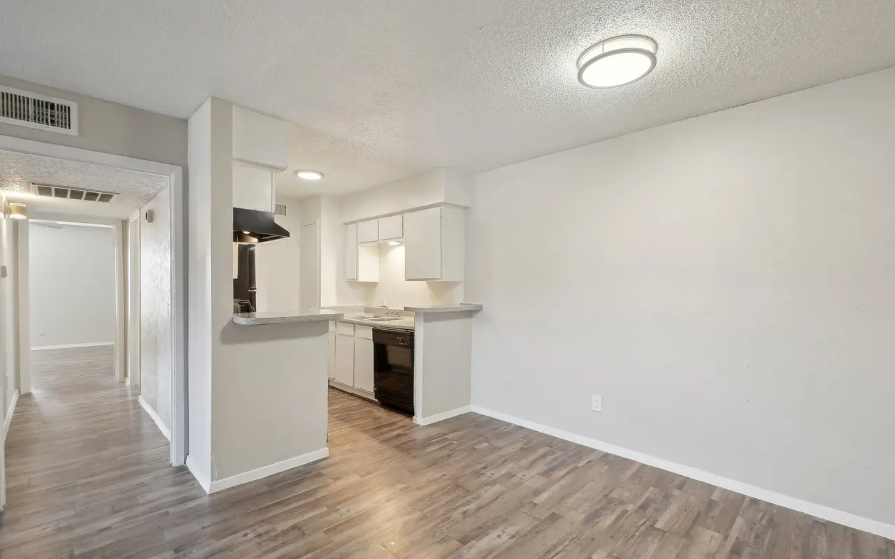 A view of a modern apartment interior featuring a kitchen with white cabinets and dark appliances, open space with wooden flooring, and a bright light fixture.