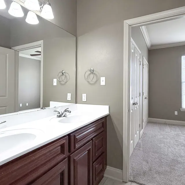 A modern bathroom featuring a double vanity with two sinks and dark wooden cabinetry. A mirror hangs above the vanity, and a doorway leads to a carpeted room with a window.