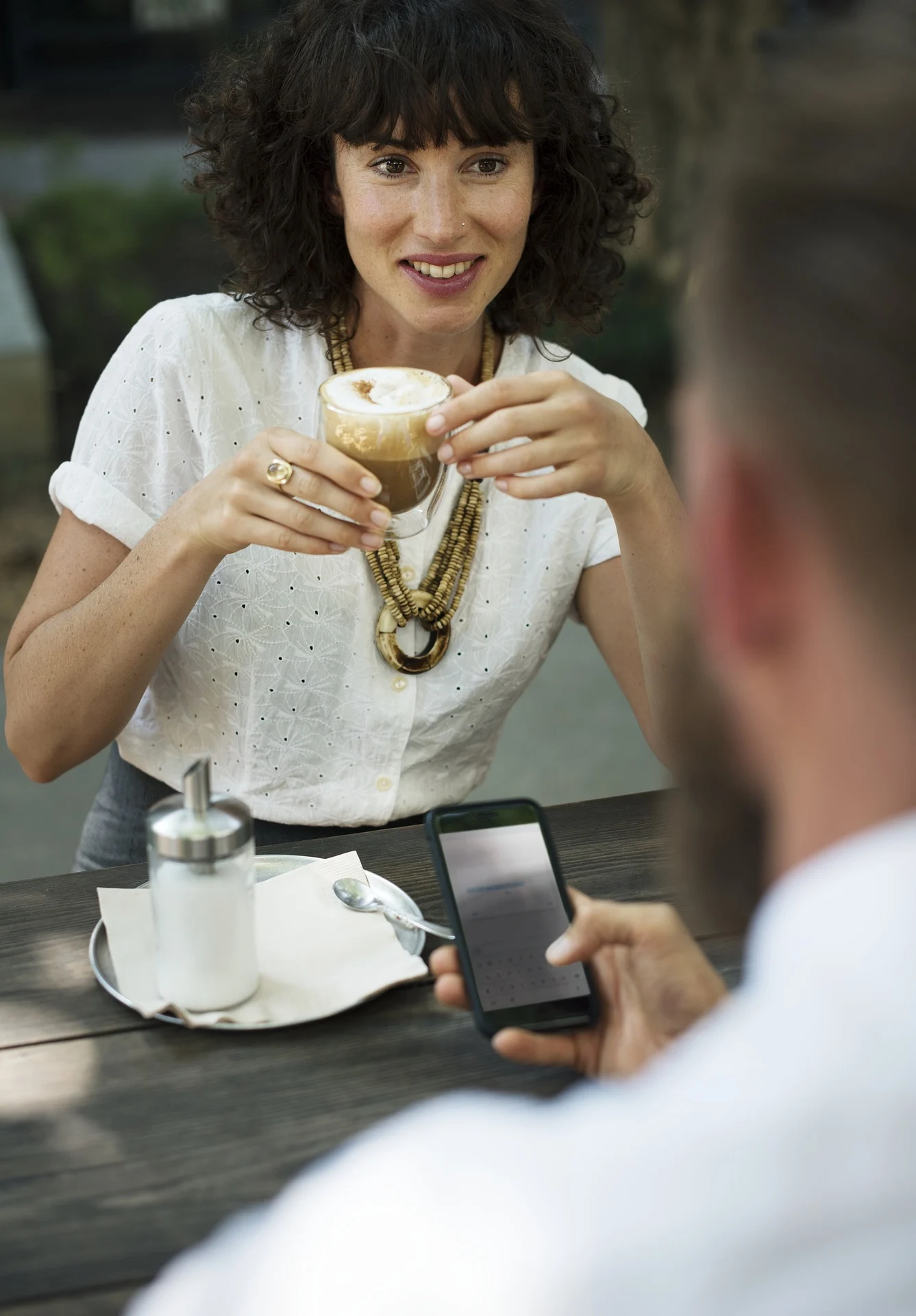 A woman with curly hair holding a coffee cup, sitting at a table with a plate and a sugar container in a cafe setting.