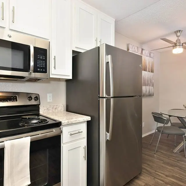 A modern kitchen featuring stainless steel appliances, including a microwave and oven, with white cabinetry, granite countertops, and a glass dining table set in the background.