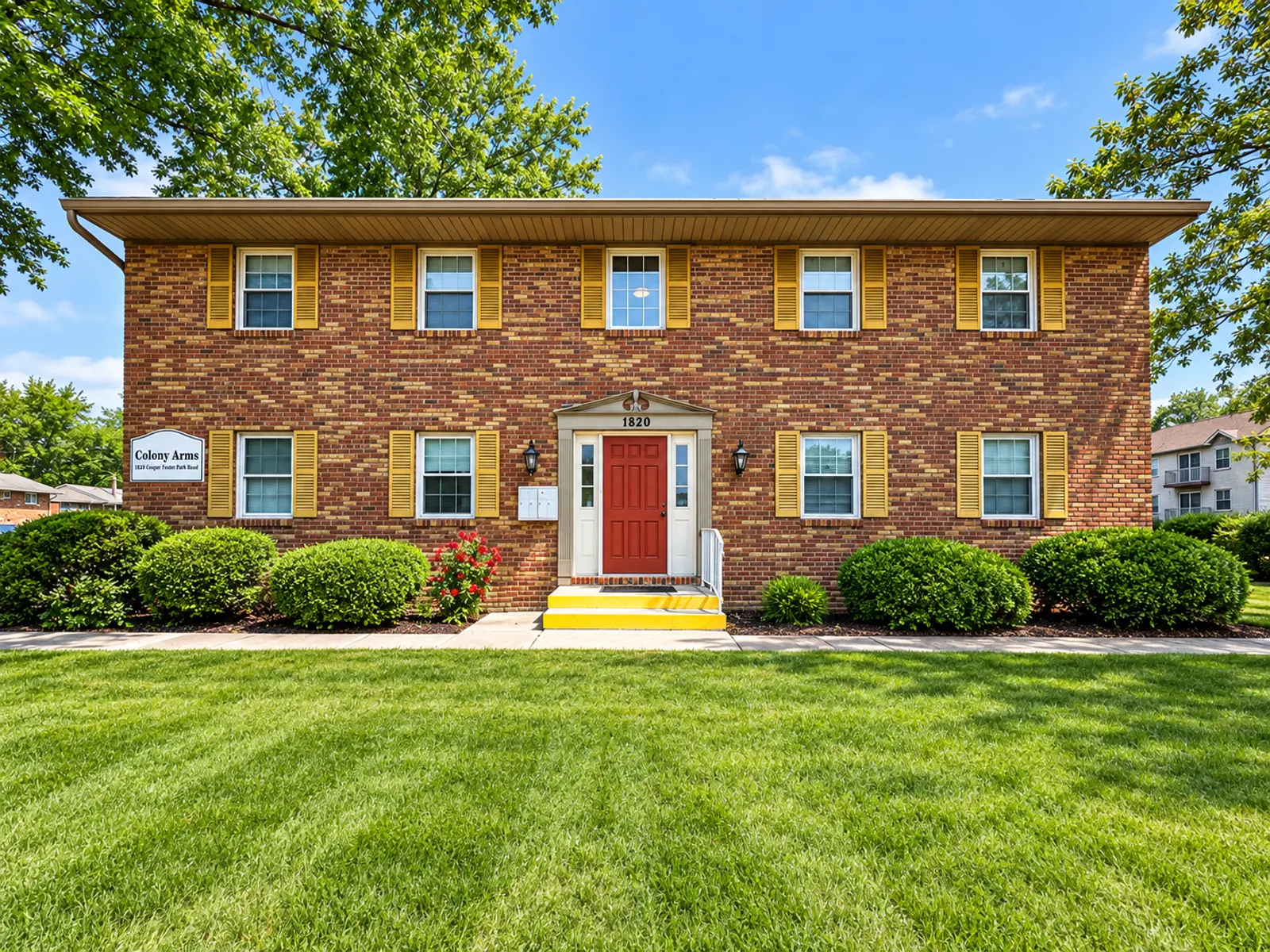 Exterior view of a two-story brick apartment building with yellow window trim and a red door, surrounded by green lawns and landscaping.