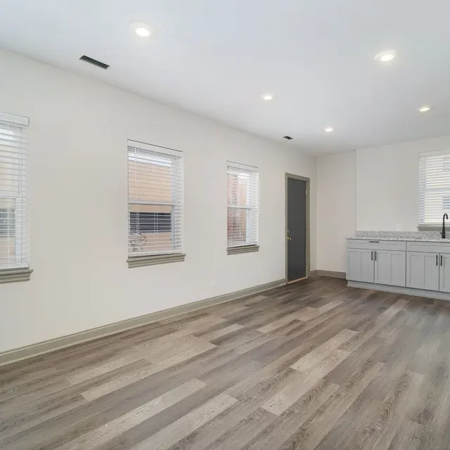 A modern apartment interior featuring light-colored walls, grey cabinetry, and large windows with white blinds. The floor is made of wood-like material and there is a kitchen area with stainless steel appliances.