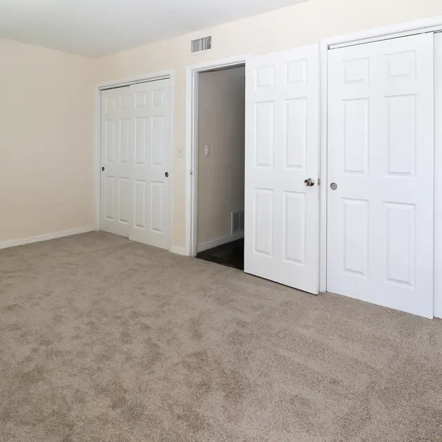 An empty room featuring light beige walls and a carpeted floor. On one side, there are two white double doors leading to closets, and on the opposite side, a door is open to another room.