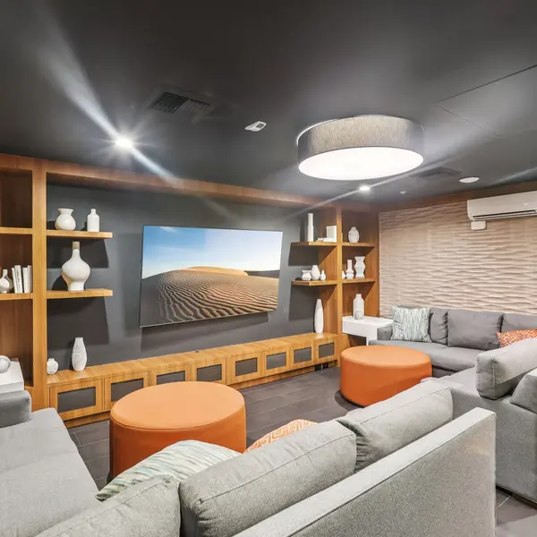 A stylish modern living room with gray sofas, orange accent tables, decorative white vases on wooden shelves, and a large wall-mounted TV displaying a desert landscape.