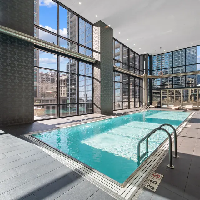 An indoor swimming pool area with large windows showcasing a city skyline. The pool is clear and inviting, surrounded by lounge chairs.