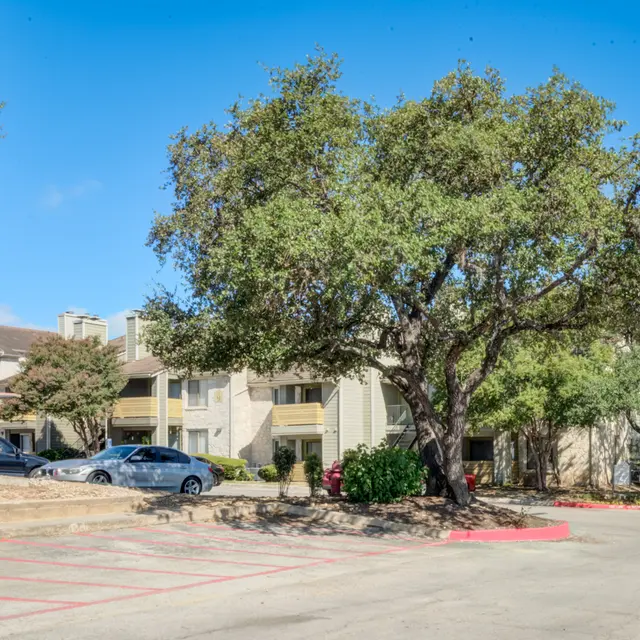 View of an apartment complex with a parking lot, featuring large trees and blue skies.