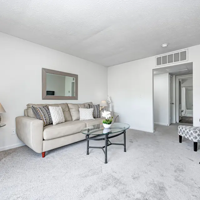 A spacious living room featuring a beige sofa with decorative pillows, a glass coffee table, and side tables with small plants. The room is carpeted, and a large mirror reflects the space. An entryway leads to another room in the background.
