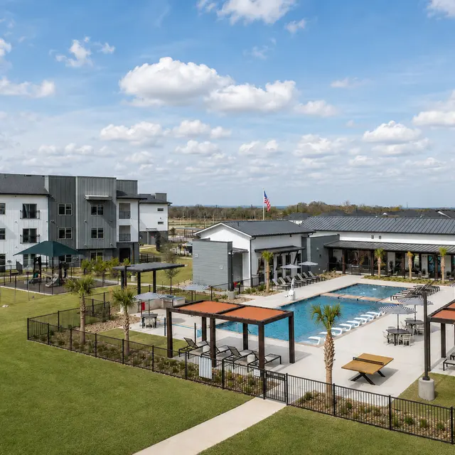 Aerial view of a luxury apartment complex featuring a swimming pool, seating areas with pergolas, and grassy open spaces under a cloudy sky.